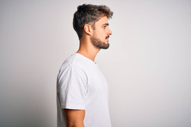 Молодой красивый мужчина с бородой носить повседневную футболку стоя на белом фоне глядя в сторону, расслабиться профиль позировать с естественным лицом с уверенной улыбкой
. - Фото, изображение