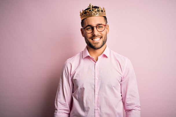 Jonge knappe zakenman draagt gouden kroon als koning over roze achtergrond met een vrolijke en koele glimlach op het gezicht. Geluksvogel. - Foto, afbeelding