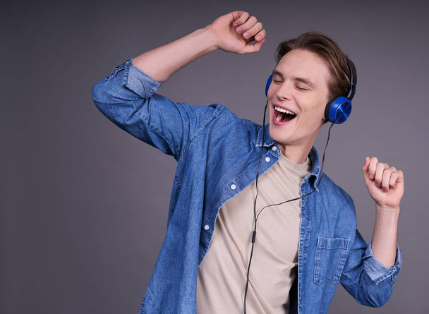 A satisfied Caucasian young man listens to a funny song in his headphones, moves against a gray background, cheers up with cool music, feels cheerful, wears a denim shirt. Have fun in your free time. - Foto, Bild