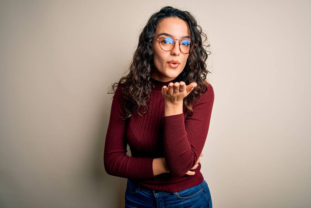 Beautiful woman with curly hair wearing casual sweater and glasses over white background looking at the camera blowing a kiss with hand on air being lovely and sexy. Love expression. - Foto, afbeelding
