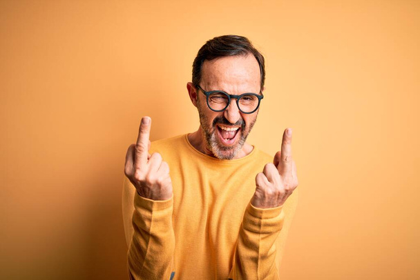 Middle age hoary man wearing casual sweater and glasses over isolated yellow background Showing middle finger doing fuck you bad expression, provocation and rude attitude. Screaming excited - Photo, Image