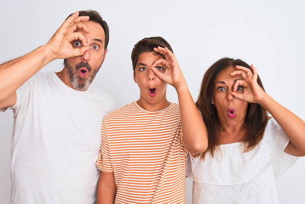 Family of three, mother, father and son standing over white isolated background doing ok gesture shocked with surprised face, eye looking through fingers. Unbelieving expression. - Foto, Bild