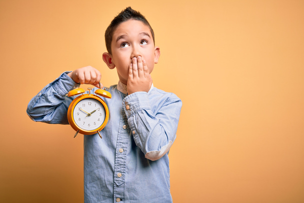 Young little boy kid holding classic bell alarm clock over isolated yellow background cover mouth with hand shocked with shame for mistake, expression of fear, scared in silence, secret concept - Foto, afbeelding