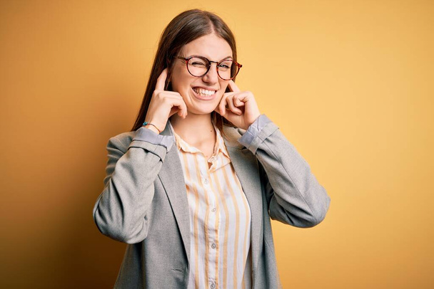 Νεαρή όμορφη κοκκινομάλλα γυναίκα φορώντας σακάκι και γυαλιά πάνω από απομονωμένο κίτρινο φόντο που καλύπτει τα αυτιά με τα δάχτυλα με ενοχλημένη έκφραση για το θόρυβο της δυνατής μουσικής. Κωφών έννοια. - Φωτογραφία, εικόνα