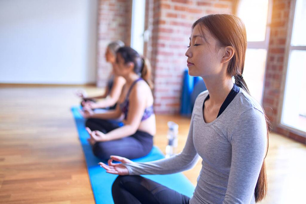 Young beautiful group of sportswomen practicing yoga. Doing lotus pose at gym - 写真・画像