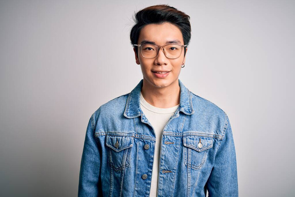 Молодой красивый китайский мужчина в джинсовой куртке и очках на белом фоне со счастливой и прохладной улыбкой на лице. Счастливчик
. - Фото, изображение