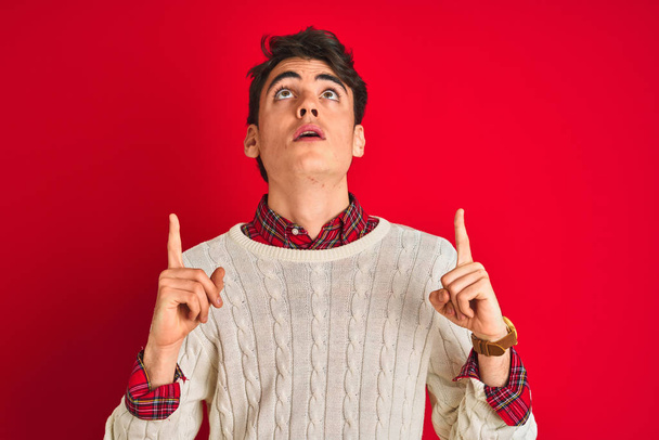 Teenager boy wearing a sweater standing over pink isolated background amazed and surprised looking up and pointing with fingers and raised arms. - Photo, Image