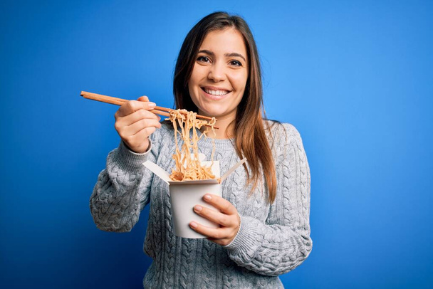 Młoda kobieta jedząca azjatycki makaron z pudełka przy użyciu pałeczki na niebieskim tle ze szczęśliwą twarzą stojącą i uśmiechniętą z pewnym uśmiechem pokazującym zęby - Zdjęcie, obraz