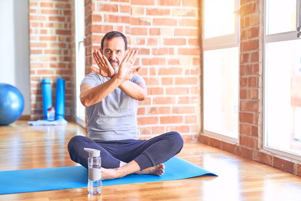 Middelbare leeftijd knappe sportman zittend op mat doen stretching yoga oefening in de sportschool Afwijzing expressie kruising armen en palmen doen negatief teken, boos gezicht - Foto, afbeelding