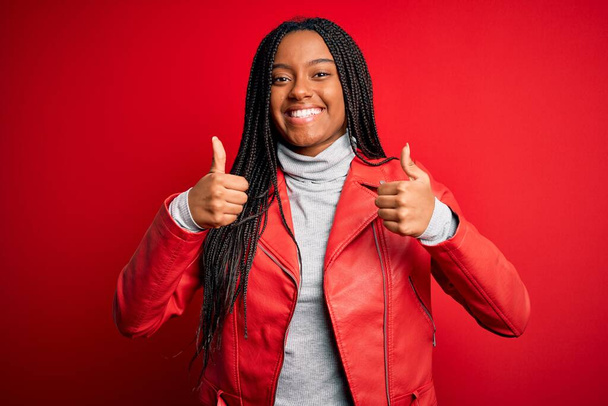 Jeune femme afro-américaine portant une veste en cuir de mode cool sur fond rouge isolé signe de succès faisant geste positif avec la main, pouces levés souriant et heureux. Expression joyeuse et geste gagnant
. - Photo, image