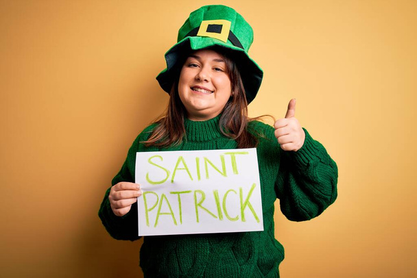 Plus Size Frau mit grünem Hut, die den Patricks Day feiert und ein Banner mit dem Heiligen Patricks Day hält, glücklich mit einem breiten Lächeln, das ok Zeichen macht, Daumen hoch mit den Fingern, hervorragendes Zeichen - Foto, Bild