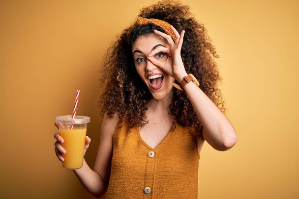 Młoda piękna kobieta z kręconymi włosami i przekłuwanie picia zdrowego soku pomarańczowego z uśmiechem szczęśliwej twarzy robi ok znak z ręki na oko patrząc przez palce - Zdjęcie, obraz
