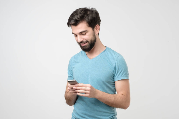 Alkalmi fiatal spanyol férfi beszél telefonon és mosolyog, miközben nézi a képernyőn. fehér alapon - Fotó, kép