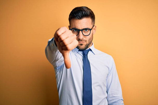 Young handsome businessman wearing tie and glasses standing over yellow background looking unhappy and angry showing rejection and negative with thumbs down gesture. Bad expression. - Photo, Image