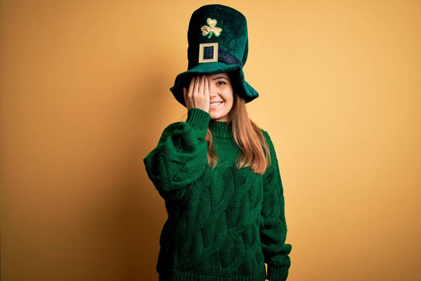 Jovem bela mulher morena usando chapéu verde em st patricks celebração do dia cobrindo um olho com a mão, sorriso confiante no rosto e emoção surpresa
. - Foto, Imagem