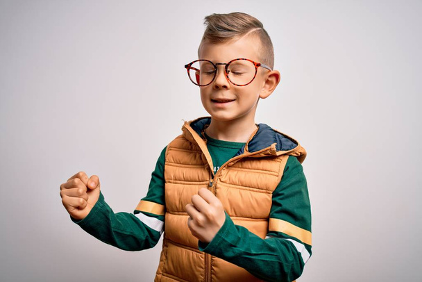 Ein kleines kaukasisches Kind mit blauen Augen, Wintermantel und smarter Brille, sehr glücklich und aufgeregt und macht Siegergeste mit erhobenen Armen, lächelt und schreit nach Erfolg. Festkonzept. - Foto, Bild