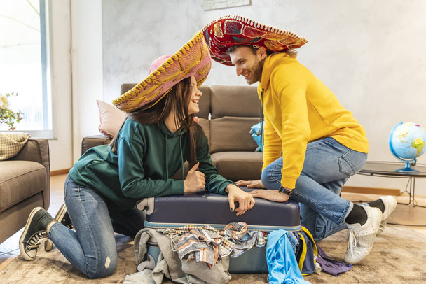 Sombrero giyen genç çift evdeki oturma odasında yapılacak bir sonraki gezi için valizleri hazırlıyor. Dünya çapında tatil kavramı - Fotoğraf, Görsel