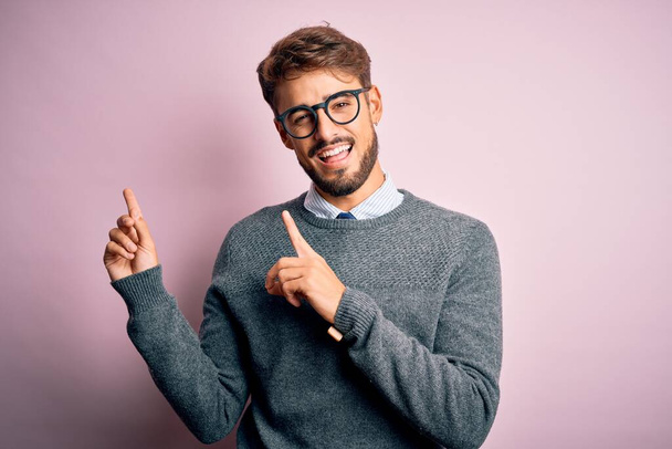 Young handsome man with beard wearing glasses and sweater standing over pink background smiling and looking at the camera pointing with two hands and fingers to the side. - Foto, afbeelding