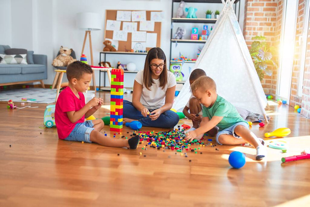 Joven hermosa maestra y niños pequeños jugando con bloques de construcción alrededor de un montón de juguetes en el jardín de infantes
 - Foto, imagen