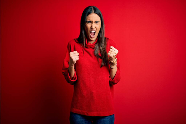 Young beautiful brunette woman wearing casual sweater over isolated red background angry and mad raising fists frustrated and furious while shouting with anger. Rage and aggressive concept. - Photo, Image