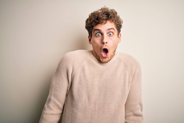 Joven hombre guapo rubio con pelo rizado usando suéter casual sobre fondo blanco asustado y sorprendido con expresión sorpresa, miedo y cara excitada
. - Foto, Imagen