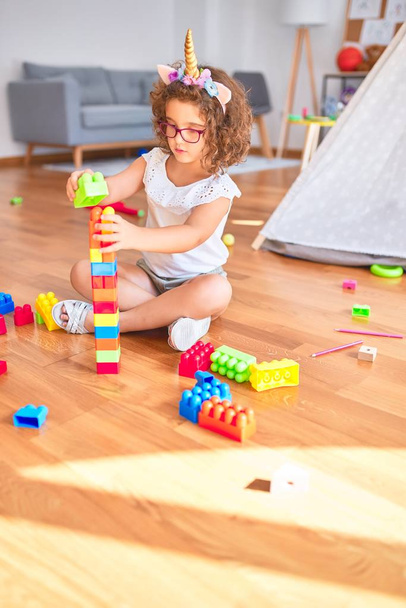 Linda criança usando óculos e diadema unicórnio sentado brincando com blocos de construção no jardim de infância
 - Foto, Imagem