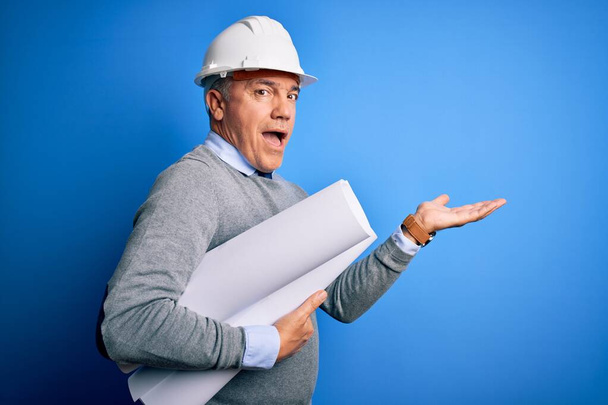 Middle age handsome grey-haired architect man wearing safety helmet holding blueprints pointing aside with hands open palms showing copy space, presenting advertisement smiling excited happy - Photo, Image