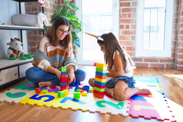 Mooie leraar en peuter spelen met bouwstenen speelgoed rond veel speelgoed op de kleuterschool - Foto, afbeelding