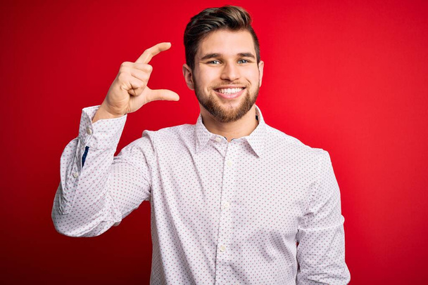 Young blond businessman with beard and blue eyes wearing elegant shirt over red background smiling and confident gesturing with hand doing small size sign with fingers looking and the camera. Measure concept. - Photo, Image