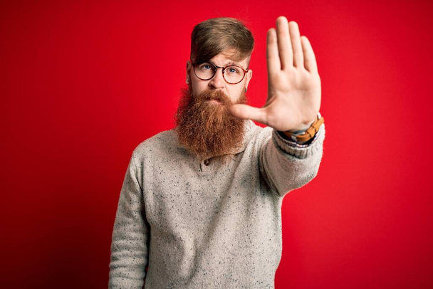 Handsome Irish redhead man with beard wearing casual sweater and glasses over red background doing stop sing with palm of the hand. Warning expression with negative and serious gesture on the face. - Photo, Image