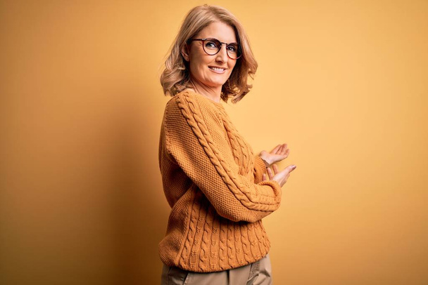 Mujer rubia hermosa de mediana edad con suéter casual y gafas sobre fondo amarillo invitando a entrar sonriendo natural con la mano abierta
 - Foto, imagen