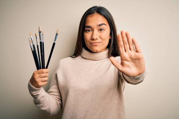 Mladý krásný umělec asijské žena drží štětce stojí nad bílým pozadím s otevřenou rukou dělat stop sign s vážným a sebejistý výraz, obrana gesto - Fotografie, Obrázek