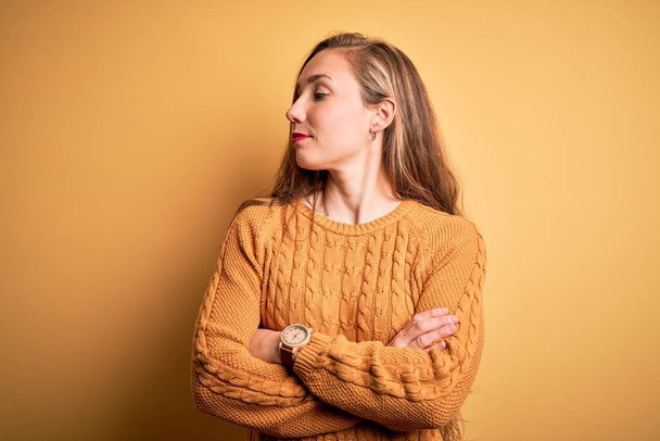 Young beautiful blonde woman wearing casual sweater standing over yellow background looking to the side with arms crossed convinced and confident - Foto, Imagen