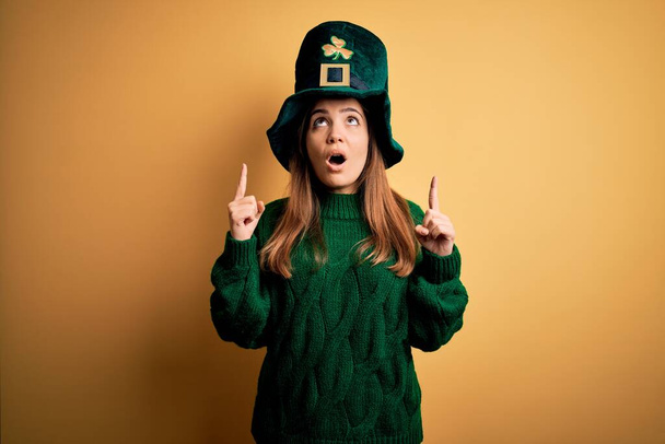 Jovem bela mulher morena usando chapéu verde em st patricks celebração do dia espantado e surpreso olhando para cima e apontando com os dedos e braços levantados
. - Foto, Imagem