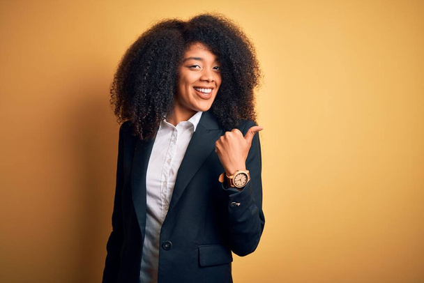 Young beautiful african american business woman with afro hair wearing elegant jacket smiling with happy face looking and pointing to the side with thumb up. - Фото, зображення
