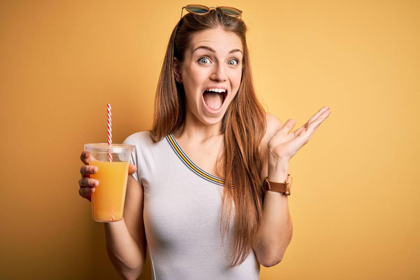 Young beautiful redhead woman drinking healthy orange juice over yellow background very happy and excited, winner expression celebrating victory screaming with big smile and raised hands - Foto, afbeelding