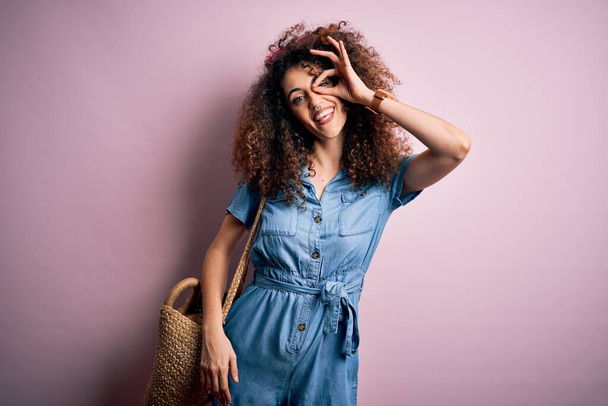 Young beautiful woman with curly hair and piercing wearing denim dress and wicker bag doing ok gesture with hand smiling, eye looking through fingers with happy face. - Photo, Image
