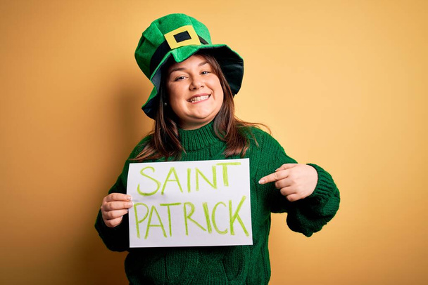 Mujer de talla grande con sombrero verde celebrando el día de San Patricio sosteniendo el estandarte del día de San Patricio muy feliz señalando con la mano y el dedo
 - Foto, imagen