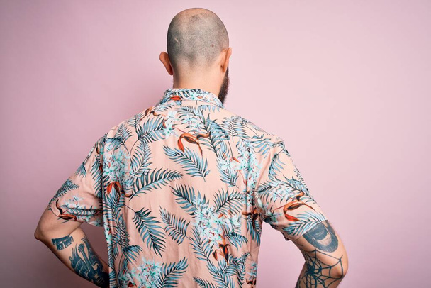 Handsome bald man with beard and tattoo wearing casual floral shirt over pink background standing backwards looking away with arms on body - Photo, Image