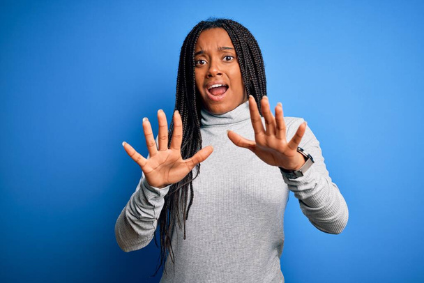 Молода афроамериканська жінка, що стоїть у звичайній білизні над синім фоном, боїться і перестає жестикулювати руками, кричачи від шоку. Панічна концепція. - Фото, зображення