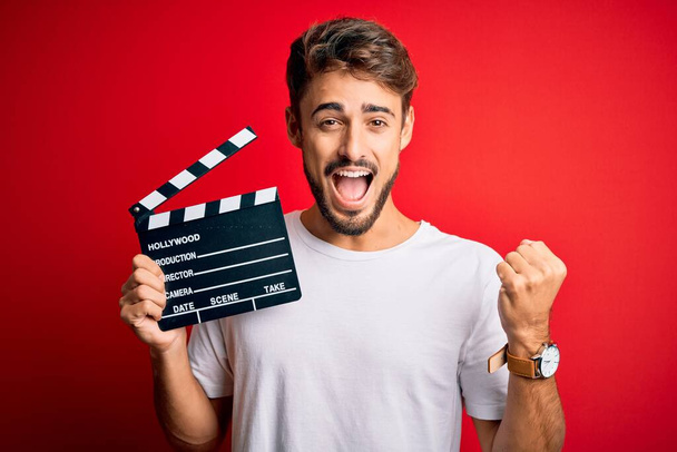 Nuori ohjaaja mies parta tehdä elokuvaa käyttäen liitutaulu yli eristetty punainen tausta huutaa ylpeä ja juhlii voittoa ja menestystä hyvin innoissaan, hurraavat tunteet
 - Valokuva, kuva