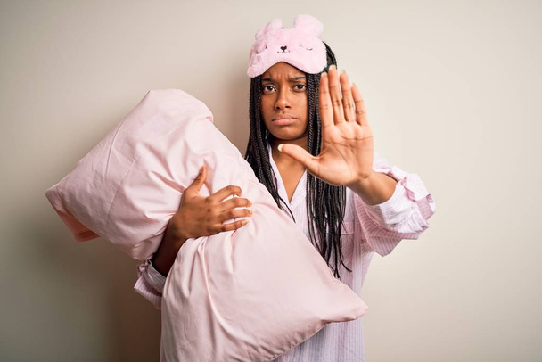 Mujer afroamericana joven con pijama y máscara de ojos abrazando almohada de la cama con la mano abierta haciendo stop sign con expresión seria y segura, gesto de defensa
 - Foto, imagen