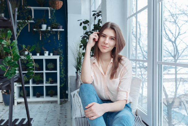 A brunette in blue jeans and a pink blouse smiles prettily and adjusts her hair. The girl sits at the window and looks at the viewer. - Фото, изображение