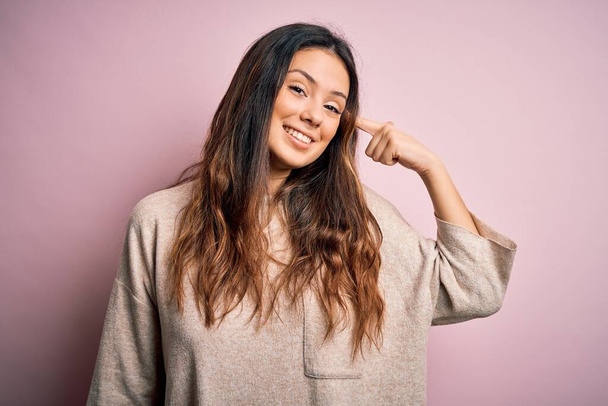 Mladá krásná brunetka žena na sobě ležérní svetr stojící nad růžovým pozadím s úsměvem ukazující na hlavu s jedním prstem, skvělý nápad nebo myšlenka, dobrá paměť - Fotografie, Obrázek