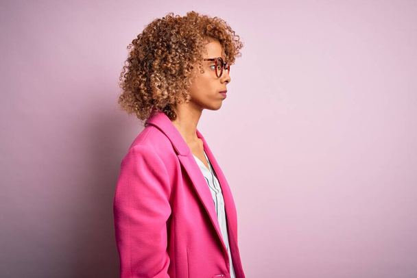 Młoda afrykańska bizneswoman nosząca okulary stojące na różowym tle patrząc na bok, relaks poza profilem z naturalną twarzą i pewny uśmiech. - Zdjęcie, obraz