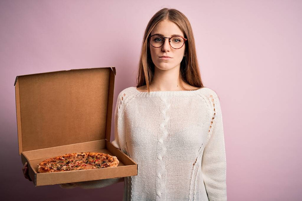 Schöne rothaarige Frau hält Lieferbox mit italienischer frischer Pizza auf rosa Hintergrund mit einem selbstbewussten Gesichtsausdruck auf intelligentem Gesichtsdenken ernst - Foto, Bild