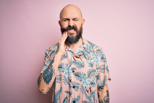 Красивый лысый мужчина с бородой и татуировкой носит случайную цветочную рубашку на розовом фоне касаясь рта с болезненным выражением лица из-за зубной боли или зубной болезни на зубах. Дантист
 - Фото, изображение