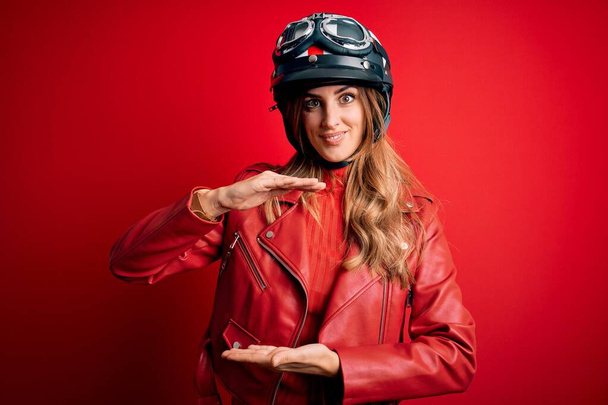 Νεαρή όμορφη μελαχρινή motrocyclist γυναίκα φορώντας moto κράνος πάνω από το κόκκινο φόντο gesturing με τα χέρια που δείχνουν μεγάλο και μεγάλο μέγεθος σημάδι, μέτρο σύμβολο. Χαμογελώντας κοιτάζοντας την κάμερα. Έννοια μέτρησης. - Φωτογραφία, εικόνα