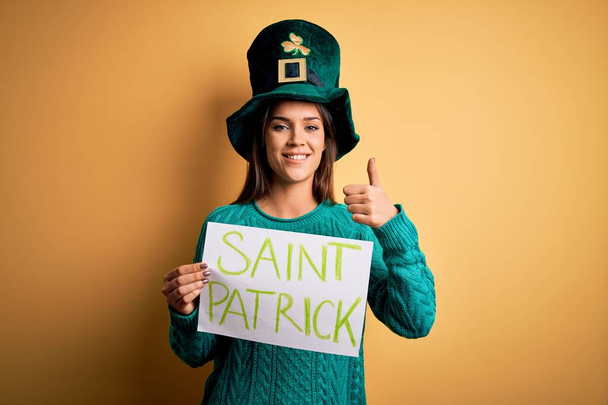 Zöld kalapos nő ünnepli a Szent Patrik napot, kezében banner szent Patrick üzenettel boldog nagy mosollyal csinál oké jel, hüvelykujj fel ujjakkal, kiváló jel - Fotó, kép