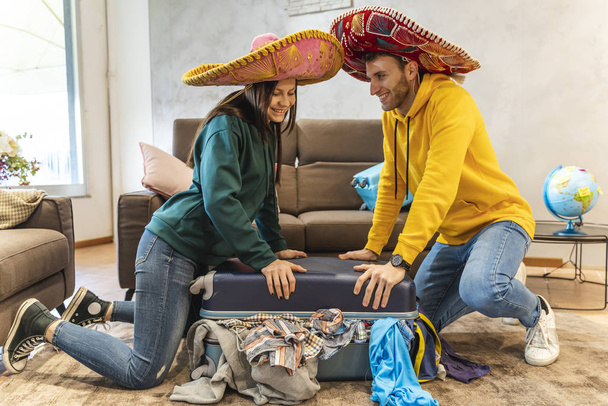 young couple wearing sombrero is preparing the suitcases for the next trip in the living room at home. holidays around the world concept - Fotoğraf, Görsel